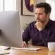 Man seated at a desk looking at a computer, with the back of the monitor facing the camera.