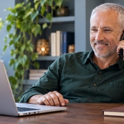 A mature man working at a laptop during a friendly phone conversation in a blue-gray office